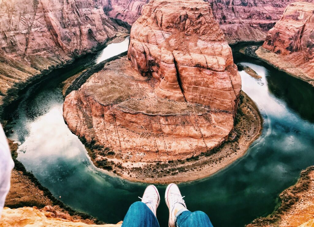 Perspektive von oben auf den Horseshoe Bend am Colorado River mit Blick über die Füße einer Person, die am Canyonrand sitzt