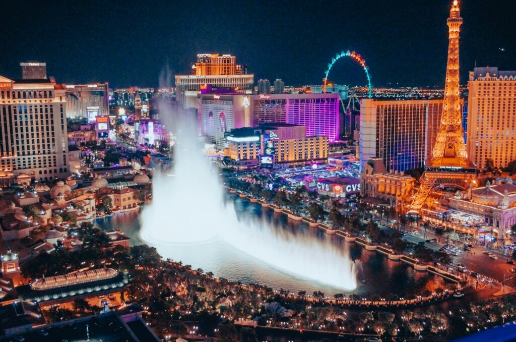 Luftaufnahme des Las Vegas Strip bei Nacht mit den beleuchteten Bellagio-Fontänen im Vordergrund und Hotels wie Paris, Bally's und Eiffelturm-Nachbau im Hintergrund